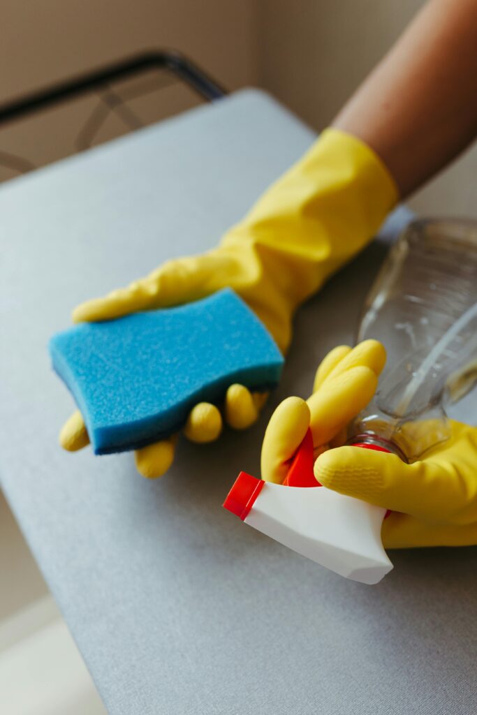 Home Close-up of a person cleaning a surface with a blue sponge and spray bottle, using yellow gloves.